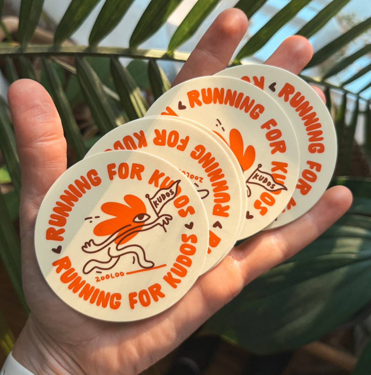 Hand holding a fan of round Running for Kudos vinyl stickers by Zooloo Design, orange flower-headed runner waving a Kudos flag on cream