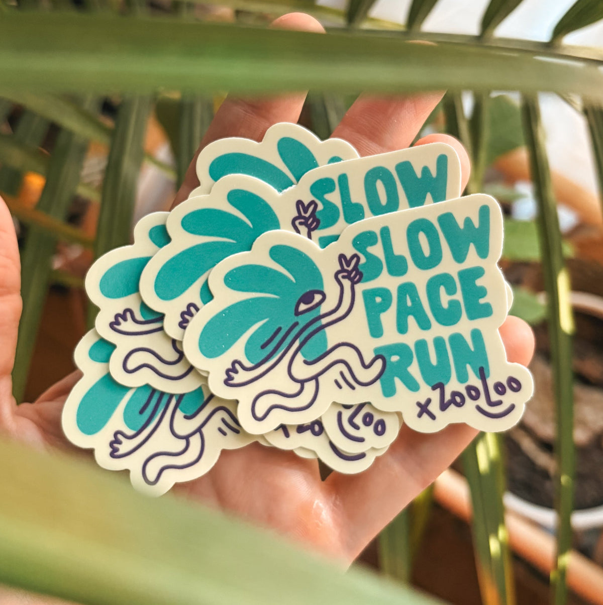 Stack of Slow Pace Run vinyl stickers by Zooloodesign, a teal palm-leaf character running and flashing a peace sign, held in hand